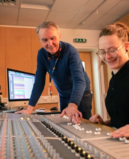 A lecturer and student working together at a large mixing console, discussing adjustments to the sound levels. A lecturer and student working together at a large mixing console, discussing adjustments to the sound levels.